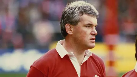 Getty Images A man with grey-brown hair wearing a red Welsh rugby jersey. He has a serious expression on his face as he looks off to the side. There's a blurry background of a rugby stadium behind him. 