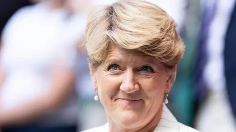 Getty Images A woman with short blonde hair wearing a white jacket and gold pearl-drop earrings. She is looking off to the side and smiling. It is a headshot. 