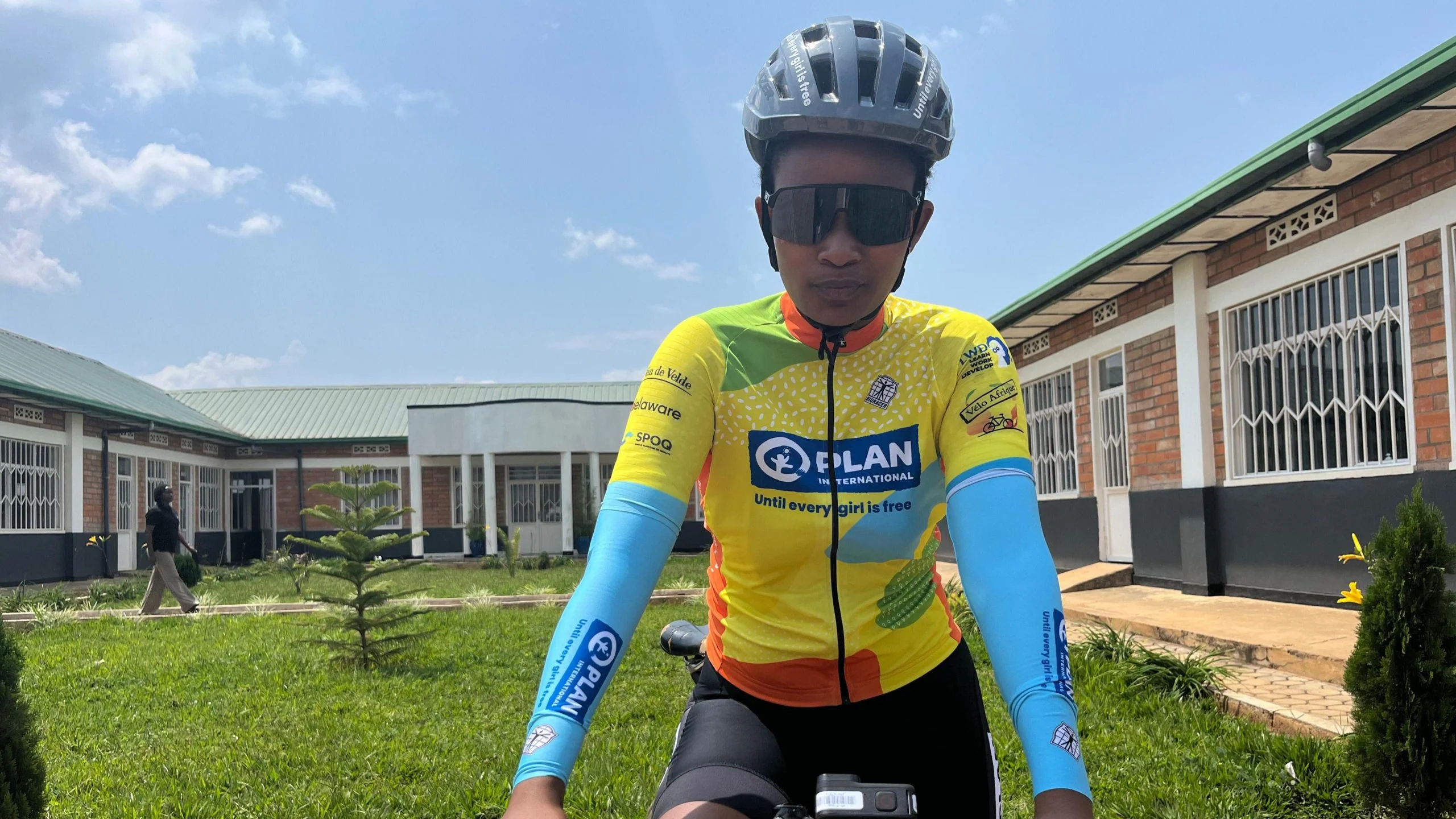 Olivia Manigragena wears a grey helmet, sunglasses and a brightly-coloured, predominantly yellow, cycling jersey as she sits on a bicycle and looks into the camera ahead of a race at the Bikes for Future centre in Bugesera. Three sides of a single-storey red brick building with white windows and a green roof can be seen in the near background behind her surrounding a patch of green grass