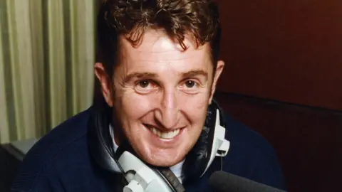 A man with curly brown haior wearing a navy fleece and smiling at the camera. He has headphones around his neck and is sat in front of a black microphone. It is a head and shoulders shot of him. 