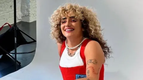 Hattie Crowther Hattie Crowther sits against a white photographic backdrop inside a studio, wearing red dungarees with an England insignia stitched onto the left side. She wears a pearl necklace and chain with spiky elements, and smiles as her blonde, curly hair cascades down her back.