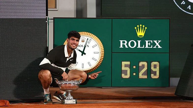 Carlos Alcaraz poses next to the clock on Court Philippe Chatrier after his five-hour and 29-minute final against Jannik Sinner