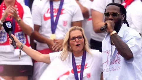 Getty Images Sarina Wiegman puts an arm around Burna Boy as he performs on-stage in front of a line of England players at the team's victory celebrations.