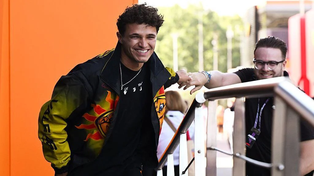 Lando Norris smiling while wearing a black and yellow jacket with a sun motif